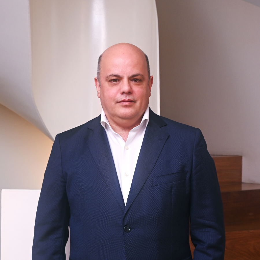 Homem de fato azul, posando em frente a escadas, num ambiente claro e moderno.