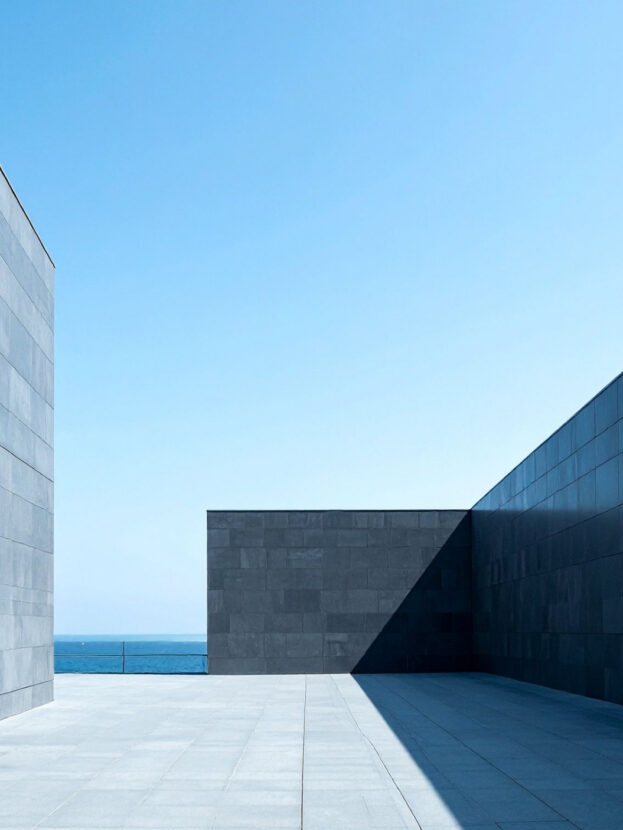Arquitetura moderna com paredes cinzas e céu azul ao fundo, estilo minimalista, junto ao mar.