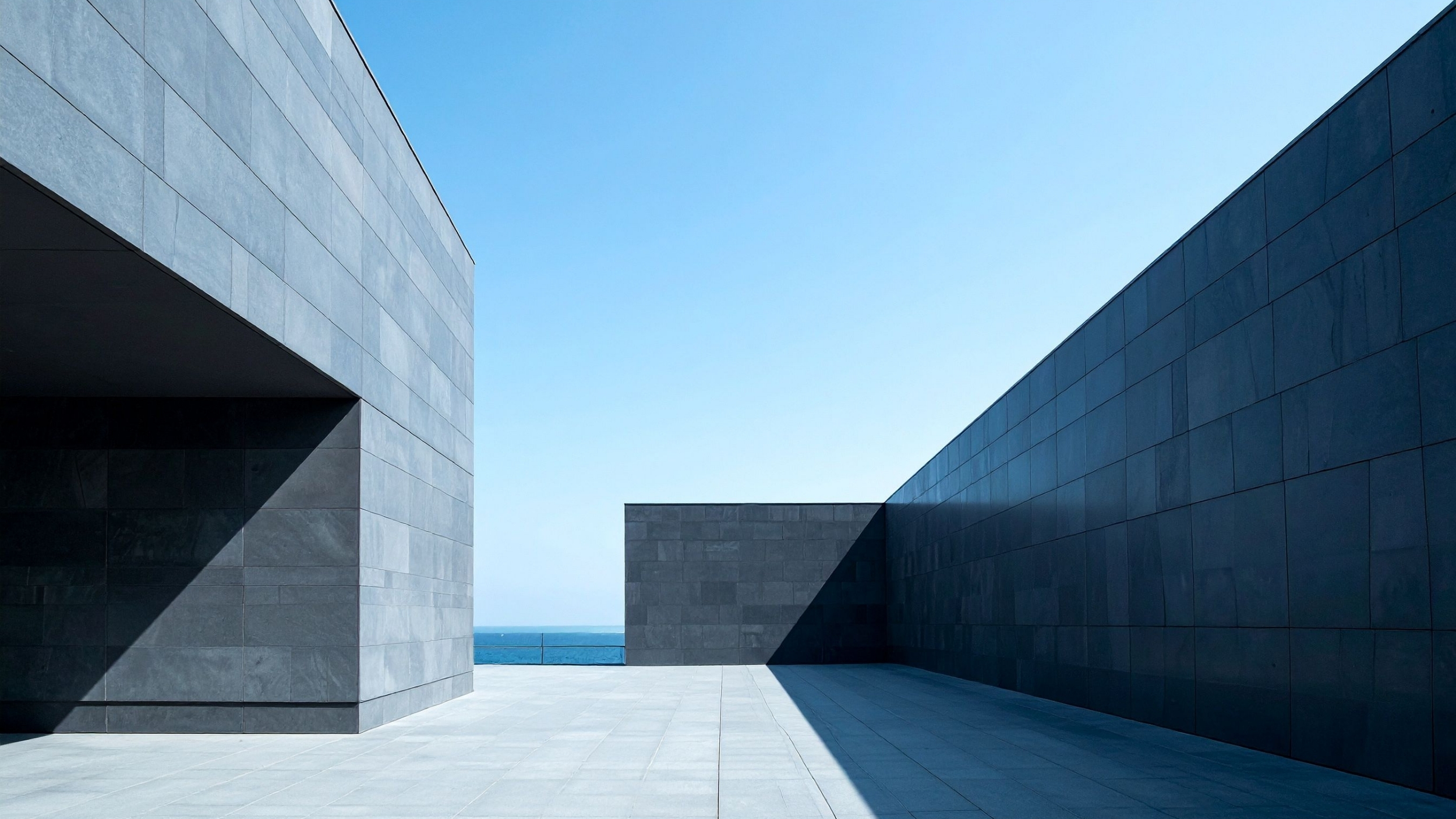 Arquitetura moderna com paredes cinzas e céu azul ao fundo, estilo minimalista, junto ao mar.