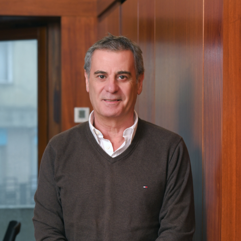 Man in a brown sweater standing indoors with wooden panel background.
