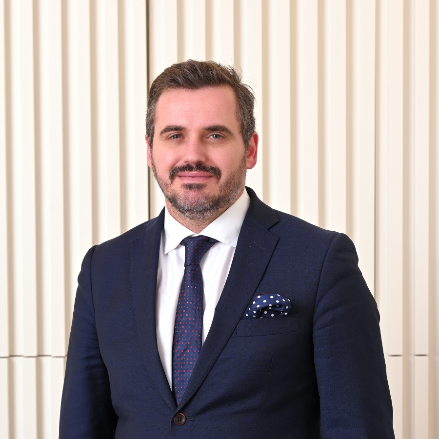 Homem de fato e gravata, com expressão profissional, em frente a fundo neutro listrado.